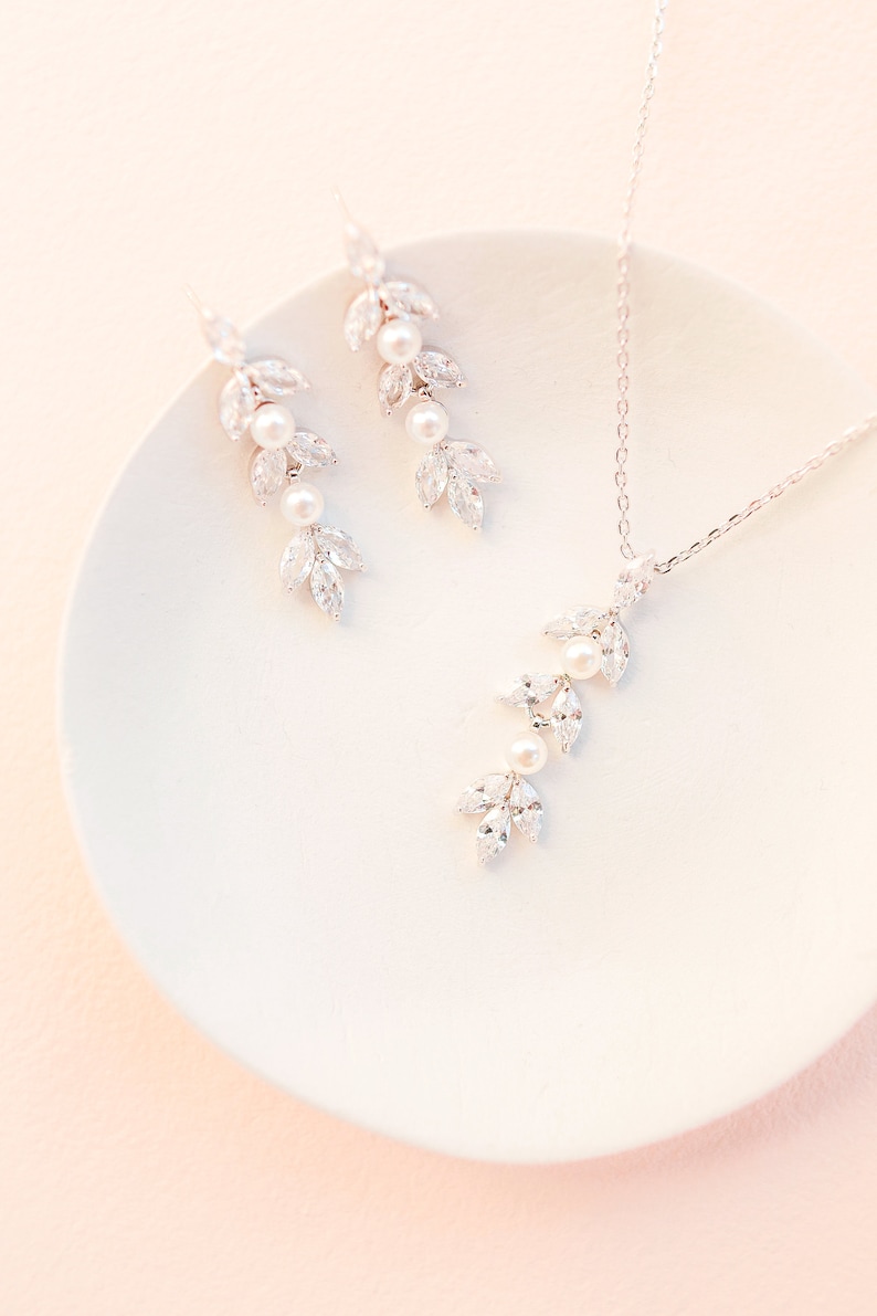 May include: A delicate jewelry set featuring a necklace and earrings. The set includes a silver-toned chain necklace and matching earrings, each adorned with leaf-shaped crystals and pearls. The jewelry is displayed on a white plate.