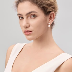 May include: A woman wearing a white sleeveless top and pearl earrings. The earrings are composed of two round pearls, one above the other. The woman has light skin, blonde hair, and is looking towards the camera.