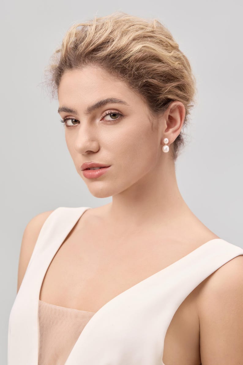 May include: A woman wearing a white sleeveless top and pearl earrings. The earrings are composed of two round pearls, one above the other. The woman has light skin, blonde hair, and is looking towards the camera.