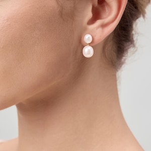 May include: A close-up of a person's ear wearing a pair of pearl earrings. Each earring features a small, round pearl above a larger, round pearl. The earrings are set on gold posts. The background is a soft, neutral color.