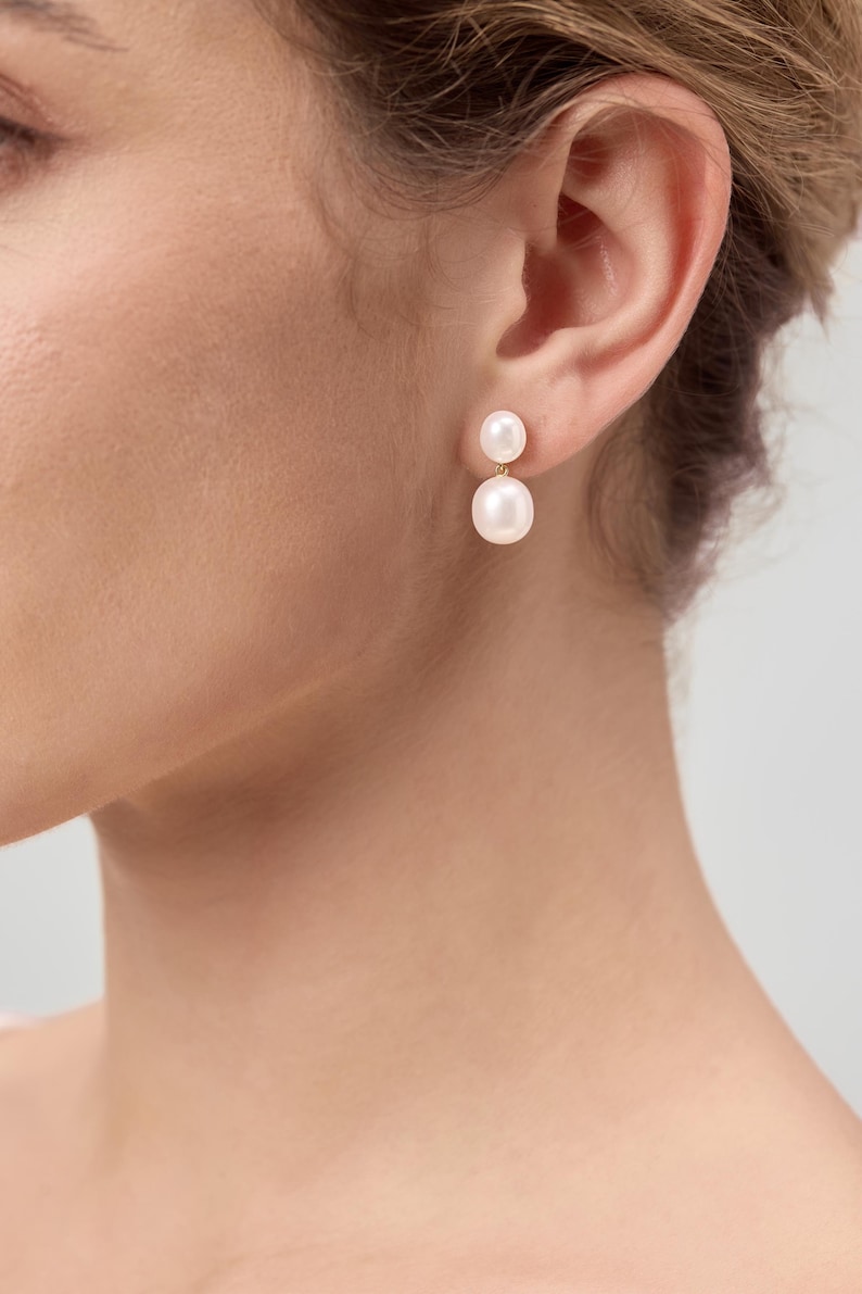 May include: A close-up of a person's ear wearing a pair of pearl earrings. Each earring features a small, round pearl above a larger, round pearl. The earrings are set on gold posts. The background is a soft, neutral color.