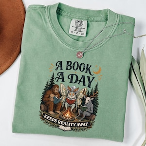 Op de afbeelding: Een saliegroen t-shirt met korte mouwen en een afbeelding van Bigfoot, een mot en een wezen met een hertenkop die boeken lezen bij een kampvuur. De tekst luidt A BOOK A DAY KEEPS REALITY AWAY. Het shirt heeft een Comfort Colors label in de nek.