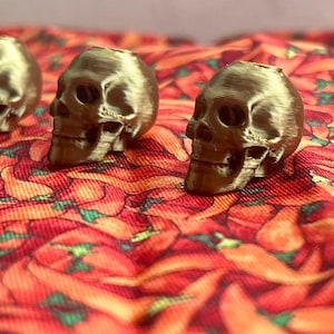 May include: Four small, gold-colored skull figurines are arranged in a row on a red fabric patterned with chili peppers. The skulls have detailed facial features and are evenly spaced, creating a striking contrast against the vibrant background.