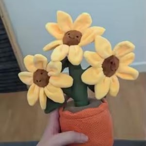 May include: A plush sunflower arrangement in an orange pot. The sunflowers have yellow petals and brown centers with smiling faces. The green stems and leaves add contrast to the bright colors. The pot is a textured orange fabric.
