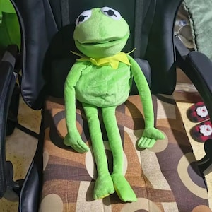 May include: A plush green frog doll with large black and white eyes, a yellow collar, and long arms and legs. The frog is sitting on a patterned chair. The doll is a popular character from a children's show.