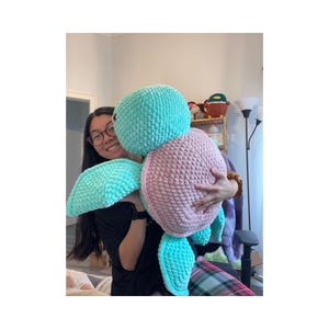 May include: A large, handmade crochet turtle plushie in shades of teal and pink. The turtle has a textured shell and flippers, and is being held by a person. The plushie is a soft, cuddly toy.