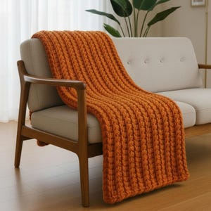 May include: An orange chunky knit throw blanket draped over a light-colored sofa and wooden chair. The blanket has a ribbed texture and adds a warm, inviting touch to the living room setting. The throw is a cozy home decor item.