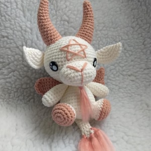 Könnte beinhalten: Gehäkeltes Ziegenplüschtier aus weißem und pfirsichfarbenem Garn. Die Ziege hat große Hörner, einen Stern auf der Stirn und kleine Flügel. Das Spielzeug hat einen flauschigen rosa Schwanz und sitzt auf einer weißen, strukturierten Oberfläche.