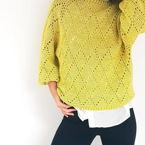 Puede incluir: Un suéter de crochet amarillo verdoso con mangas tres cuartos. El suéter tiene un patrón de diamantes y se usa sobre una camisa blanca con cuello y pantalones azul oscuro. El fondo es una pared blanca lisa.