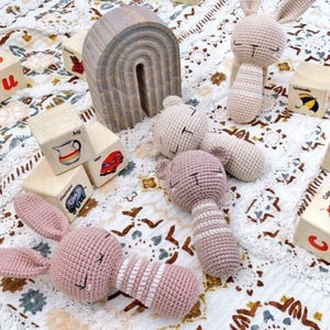 以下が含まれることがあります： ピンクとベージュのかぎ針編みのウサギのガラガラ、木製のアルファベットブロック、木製の虹など、手作りのベビーおもちゃのコレクション。おもちゃは模様入りの毛布の上に配置されています。