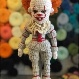 May include: A handmade crochet doll of a clown with white skin, red accents, and orange hair. The doll is wearing a gray outfit with red trim and buttons. The clown has a sinister expression.