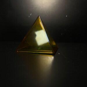 Op de afbeelding: Een transparante, goudgele piramide met een vierkante basis. De piramide is gemaakt van een glanzend materiaal en staat tegen een donkere achtergrond. Het licht reflecteert op de oppervlakken van de piramide, waardoor highlights en schaduwen ontstaan.
