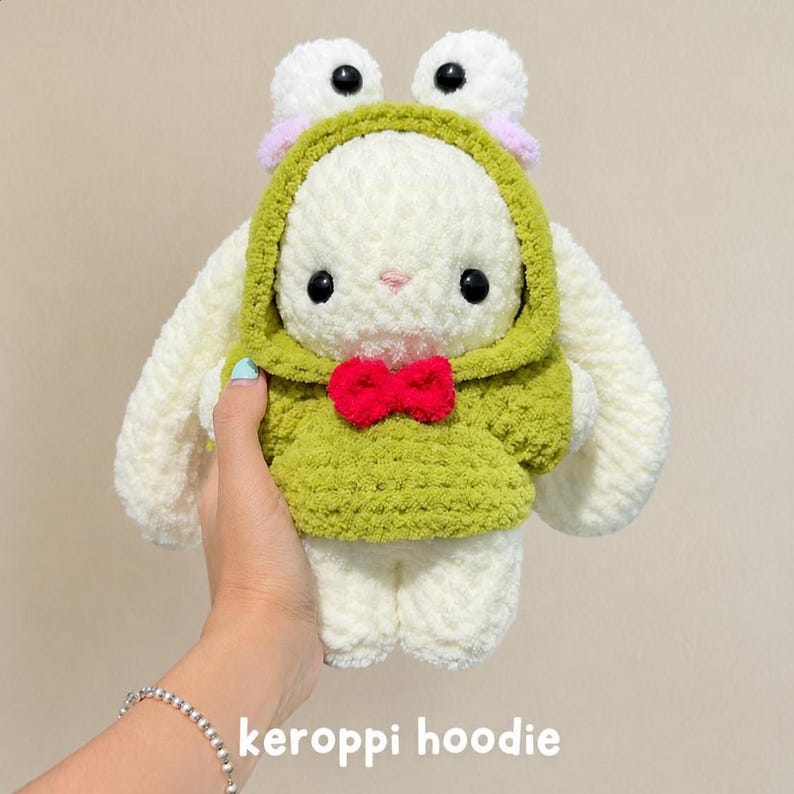 May include: A white crocheted plush toy featuring long ears and a green hooded sweatshirt with a red bow tie. The hood is designed with two frog eyes on top, each with a black pupil and a light pink accent. The text "keroppi hoodie" is visible.