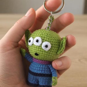 May include: A handmade crochet keychain featuring a green alien character with three eyes, small ears, and a blue outfit. The keychain has a silver metal ring for attaching to keys or bags. The alien is approximately 3 inches tall.