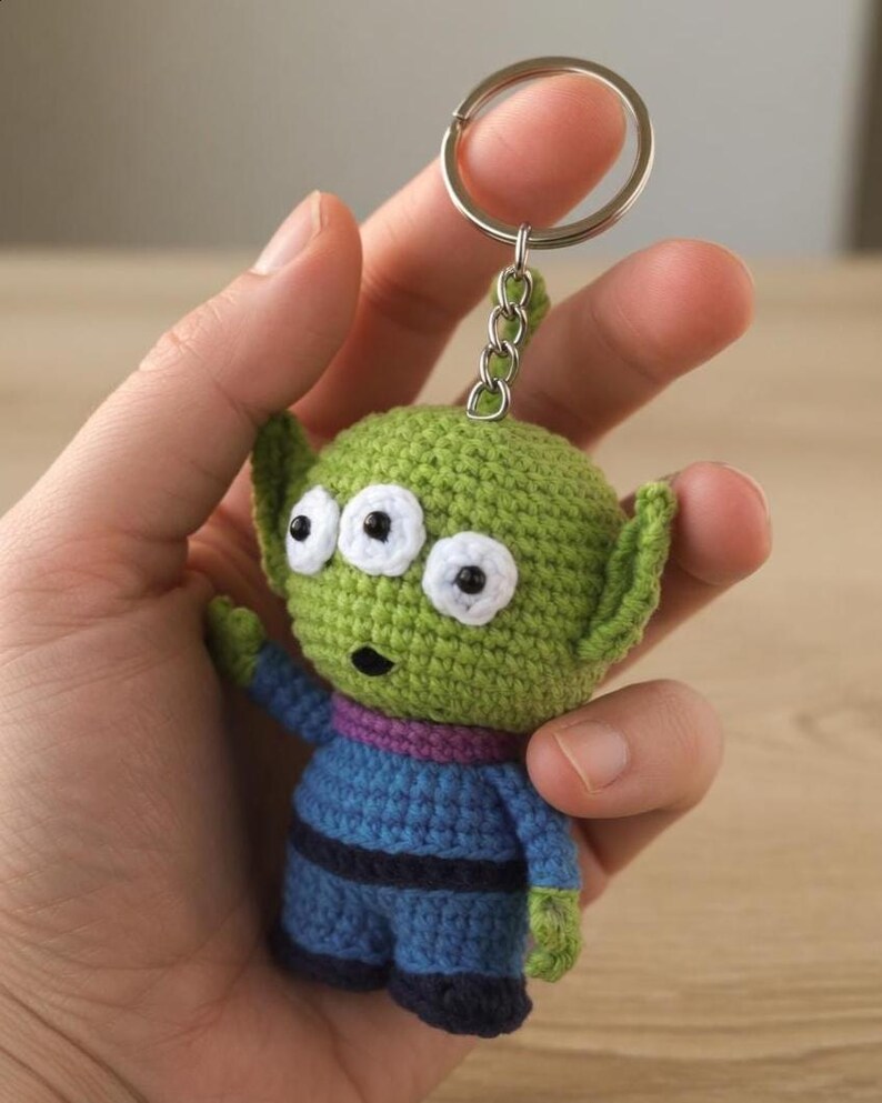 May include: A handmade crochet keychain featuring a green alien character with three eyes, small ears, and a blue outfit. The keychain has a silver metal ring for attaching to keys or bags. The alien is approximately 3 inches tall.