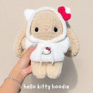 May include: A plush beige bunny toy wearing a white hooded sweatshirt. The hoodie features a small Hello Kitty graphic on the front and a bright red bow attached to the hood. The bunny has black eyes, a pink stitched nose, and long ears.