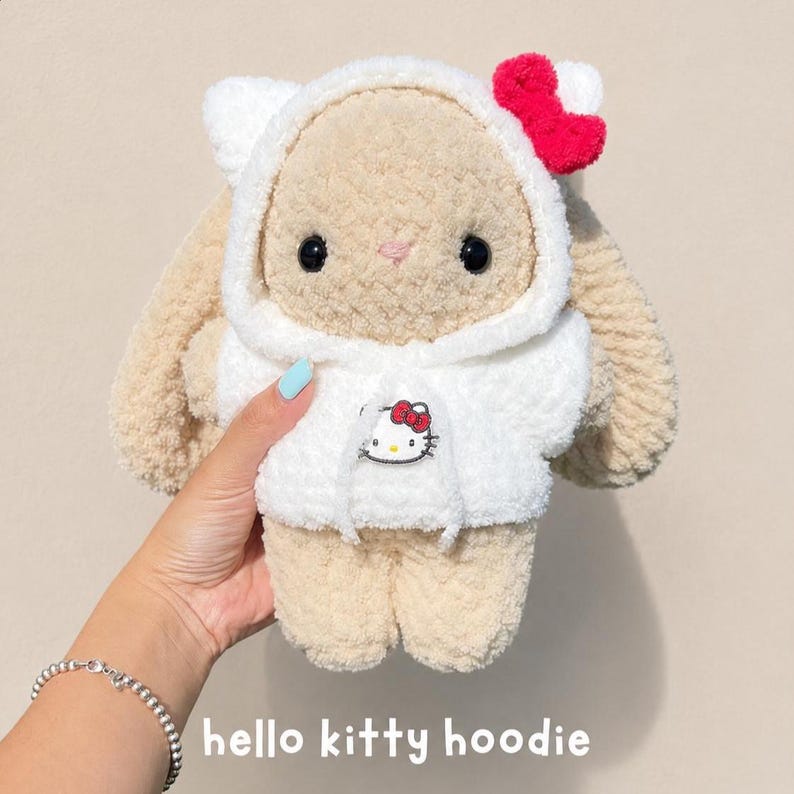 May include: A plush beige bunny toy wearing a white hooded sweatshirt. The hoodie features a small Hello Kitty graphic on the front and a bright red bow attached to the hood. The bunny has black eyes, a pink stitched nose, and long ears.