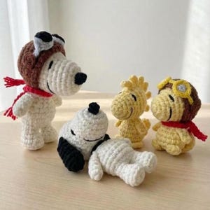 Op de afbeelding: Gehaakte Snoopy- en Woodstock-figuren. Snoopy draagt een bruine pilotenmuts en een rode sjaal. Eén Snoopy ligt. Woodstock is geel en één draagt een bruine pilotenmuts en een rode sjaal. De figuren staan op een lichtbruin oppervlak.