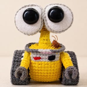 Puede incluir: Un juguete de robot de ganchillo hecho a mano en amarillo, gris y negro. El robot tiene ojos grandes y redondos con pupilas negras, un cuerpo cilíndrico y un pequeño insecto marrón en la parte superior. El juguete es una representación caprichosa de un personaje animado popular.