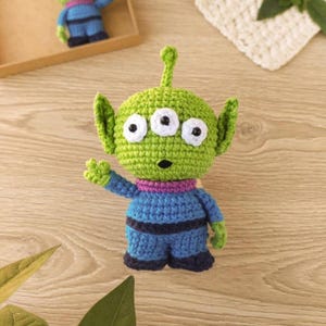 May include: A handmade green crocheted alien toy with three eyes, large ears, and a small antenna. The alien is wearing a blue outfit with purple accents. The toy is a whimsical, handcrafted item.