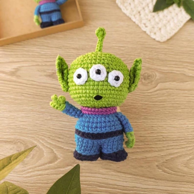 May include: A handmade green crocheted alien toy with three eyes, large ears, and a small antenna. The alien is wearing a blue outfit with purple accents. The toy is a whimsical, handcrafted item.