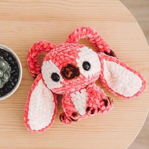 Pu&ograve; includere: Un giocattolo di peluche fatto a mano a forma di coniglietto. Il coniglietto &egrave; principalmente rosa con accenti bianchi e marroni. Ha grandi occhi neri e lunghe orecchie flosce. Il giocattolo &egrave; realizzato in un materiale morbido e strutturato, su una superficie di legno.