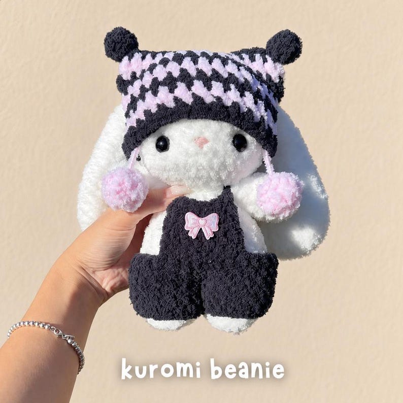 May include: A plush white rabbit toy wearing a black and light pink checkered beanie with two small pom-poms and a black pair of overalls featuring a small pink bow detail. The text kuromi beanie is printed at the bottom of the image.