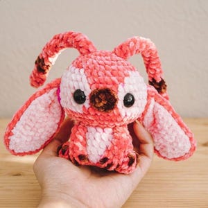 Pu&ograve; includere: Un peluche a forma di coniglietto fatto a mano all'uncinetto nei toni del rosa e del bianco. Il peluche ha grandi orecchie flosce con accenti marroni, occhi di sicurezza neri e un naso marrone. Il coniglietto &egrave; tenuto in una mano.