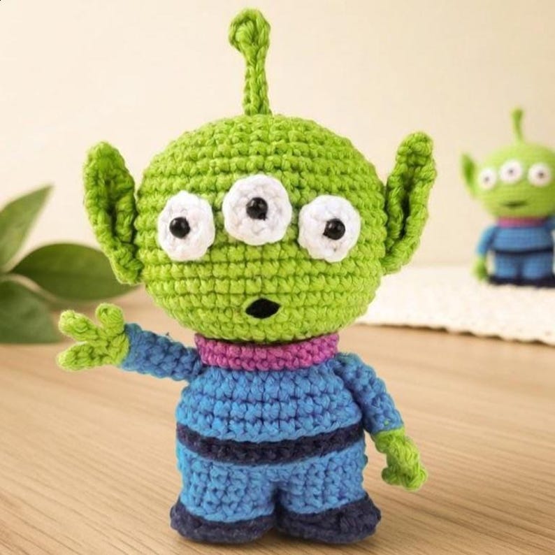 May include: A handmade green crochet alien with three white-rimmed eyes and a small antenna. The alien wears a blue outfit with a purple collar. A second, smaller alien is visible in the background.