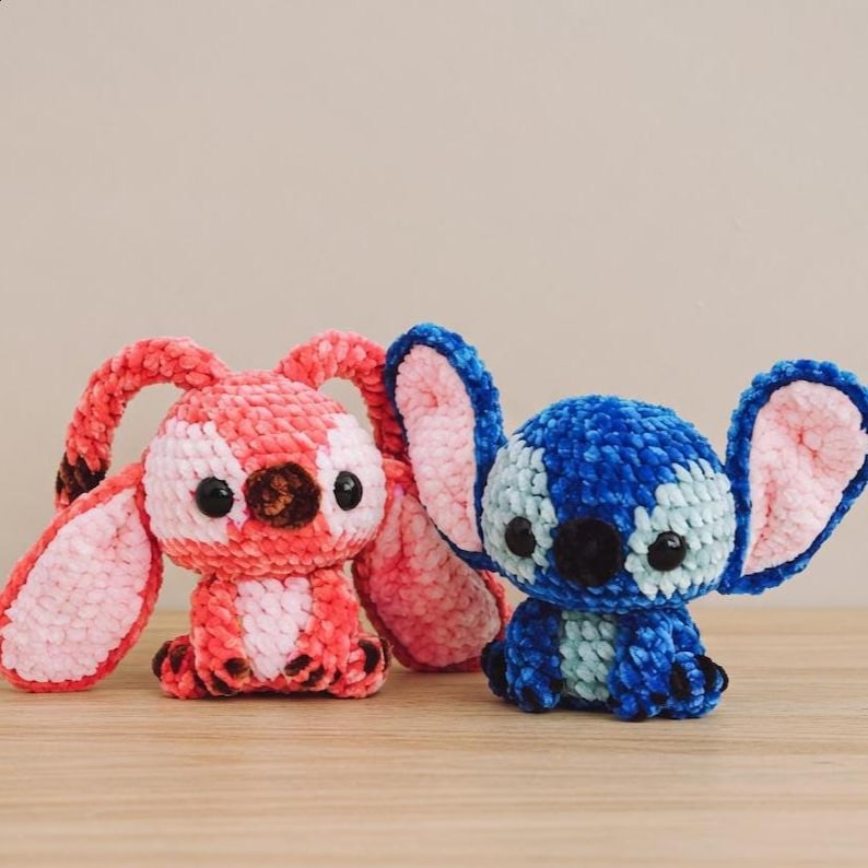 Pu&ograve; includere: Due peluche fatti a mano all'uncinetto. Uno &egrave; rosa e rosso con grandi orecchie, l'altro &egrave; blu e azzurro con grandi orecchie. Entrambi hanno occhi neri e sono su una superficie di legno.