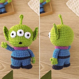May include: A handmade crochet alien toy in green, blue, and purple. The alien has three eyes, large ears, and a small antenna. It is wearing a blue jumpsuit with dark blue stripes and a purple collar. The toy is displayed on a light brown surface.