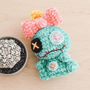 Pu&ograve; includere: Un giocattolo di peluche turchese fatto a mano con un fiocco rosa e un accento giallo. Il giocattolo ha una benda sull'occhio nera, una bocca cucita e due toppe bianche con croci rosa. Un piccolo cactus in un vaso grigio &egrave; sullo sfondo.
