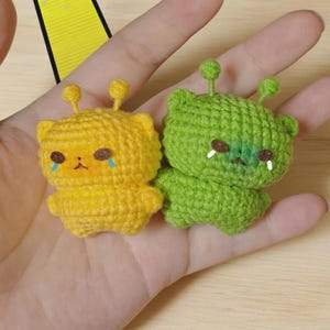 Könnte beinhalten: Zwei kleine, handgefertigte, häkelartige Kreaturen. Eine ist gelb mit blauen Tränen, die andere grün. Beide haben kleine Antennen und werden in einer Hand gehalten.