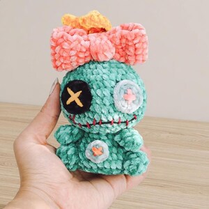 Pu&ograve; includere: Bambola di peluche turchese fatta a mano con un grande fiocco rosa e un fiocco arancione pi&ugrave; piccolo sopra. La bambola ha dettagli cuciti, tra cui un occhio nero con una croce gialla, un occhio bianco con una croce rosa e una bocca cucita rossa.