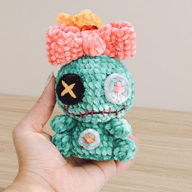 Pu&ograve; includere: Bambola di peluche turchese fatta a mano con un grande fiocco rosa e un fiocco arancione pi&ugrave; piccolo sopra. La bambola ha dettagli cuciti, tra cui un occhio nero con una croce gialla, un occhio bianco con una croce rosa e una bocca cucita rossa.