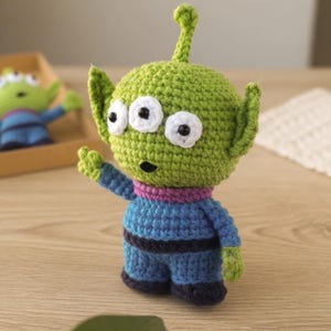 May include: A handmade green crochet alien with three white eyes and black pupils. The alien is wearing a blue outfit with a purple collar. A second, smaller alien is visible in the background.