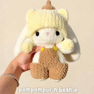 May include: A plush white rabbit toy wearing a yellow knit beanie with a brown top accent and brown overalls featuring small yellow flower buttons. The toy has black eyes and long ears. The text pompompurin beanie is visible at the bottom.