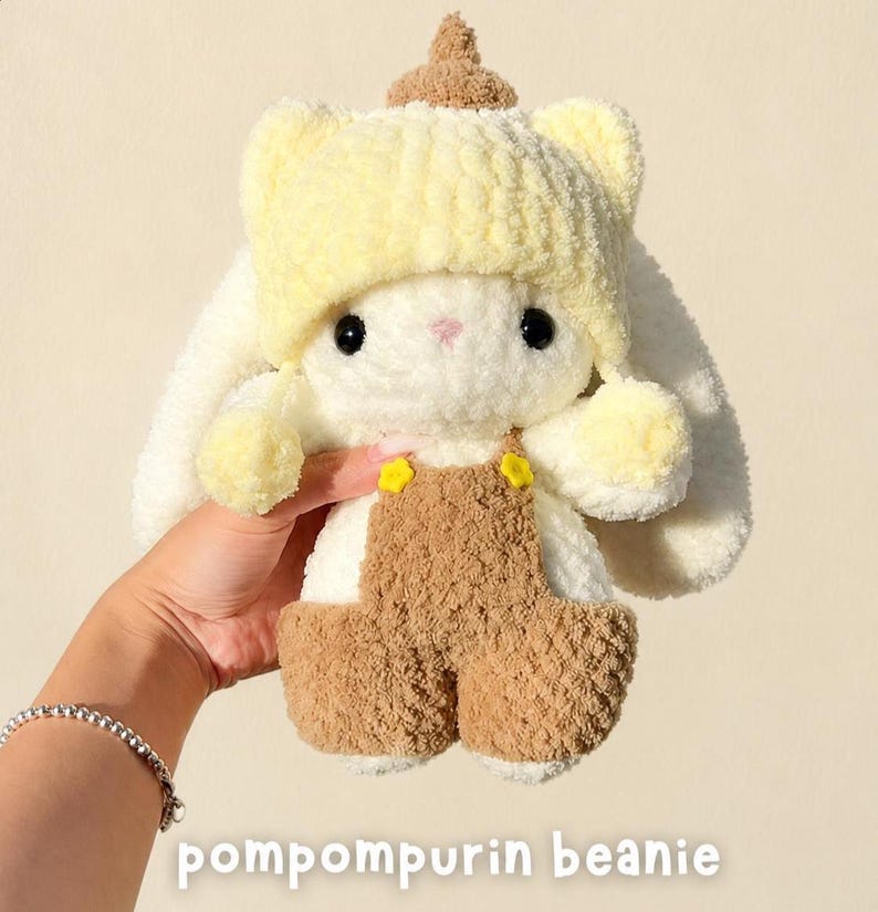 May include: A plush white rabbit toy wearing a yellow knit beanie with a brown top accent and brown overalls featuring small yellow flower buttons. The toy has black eyes and long ears. The text pompompurin beanie is visible at the bottom.