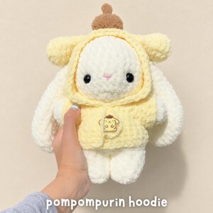 May include: A plush toy featuring a white rabbit wearing a soft yellow hoodie with a brown beret on top. The hoodie displays an embroidered Pompompurin character graphic on the front. The toy has black button eyes and a small pink stitched nose.