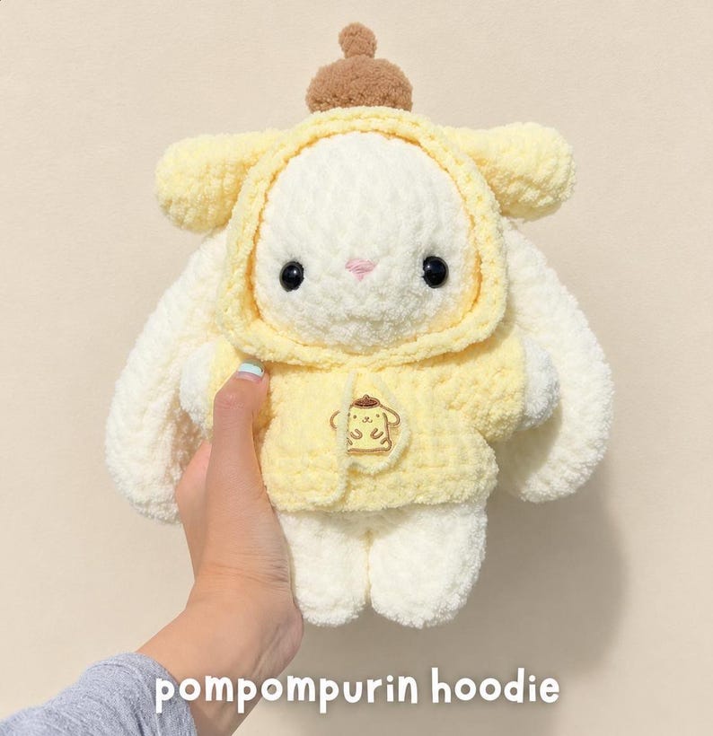 May include: A plush toy featuring a white rabbit wearing a soft yellow hoodie with a brown beret on top. The hoodie displays an embroidered Pompompurin character graphic on the front. The toy has black button eyes and a small pink stitched nose.