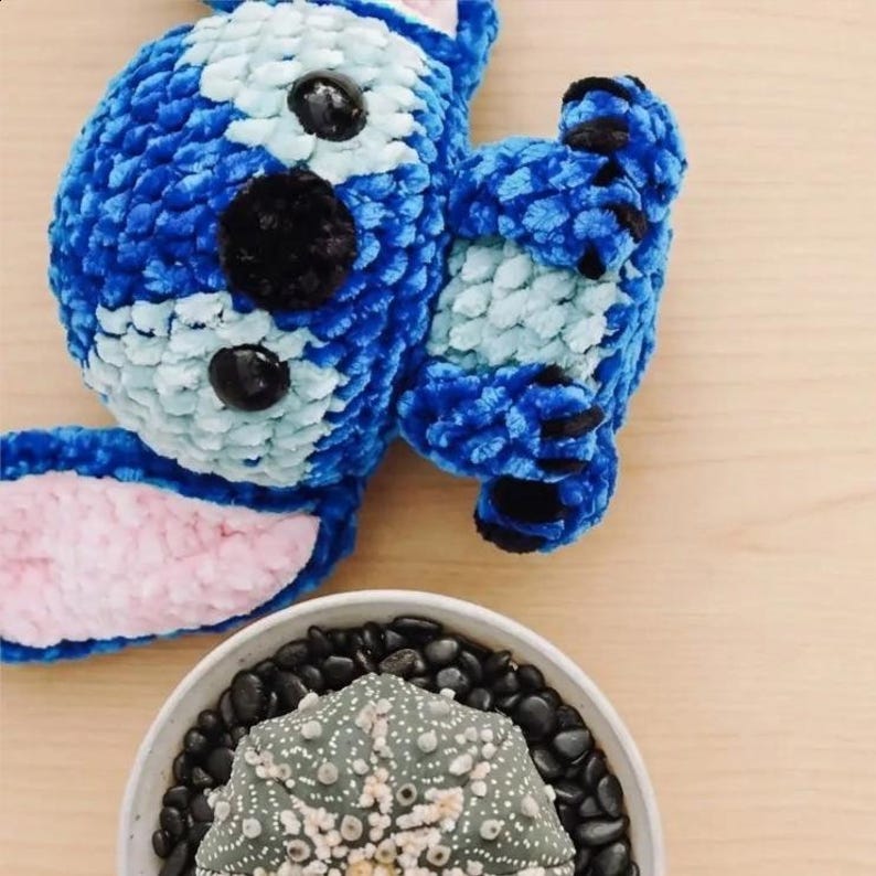 Pu&ograve; includere: Un personaggio all'uncinetto blu e bianco fatto a mano con dettagli neri e pancia rosa. Una piccola ciotola di ceramica bianca piena di pietre nere e un cactus verde in primo piano.
