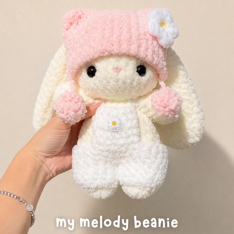 May include: A plush cream-colored rabbit toy wearing a soft pink knit beanie with a white flower accent and two pink pom-poms. The rabbit is dressed in white crocheted overalls featuring a small white flower on the chest. Text reads: my melody beanie.