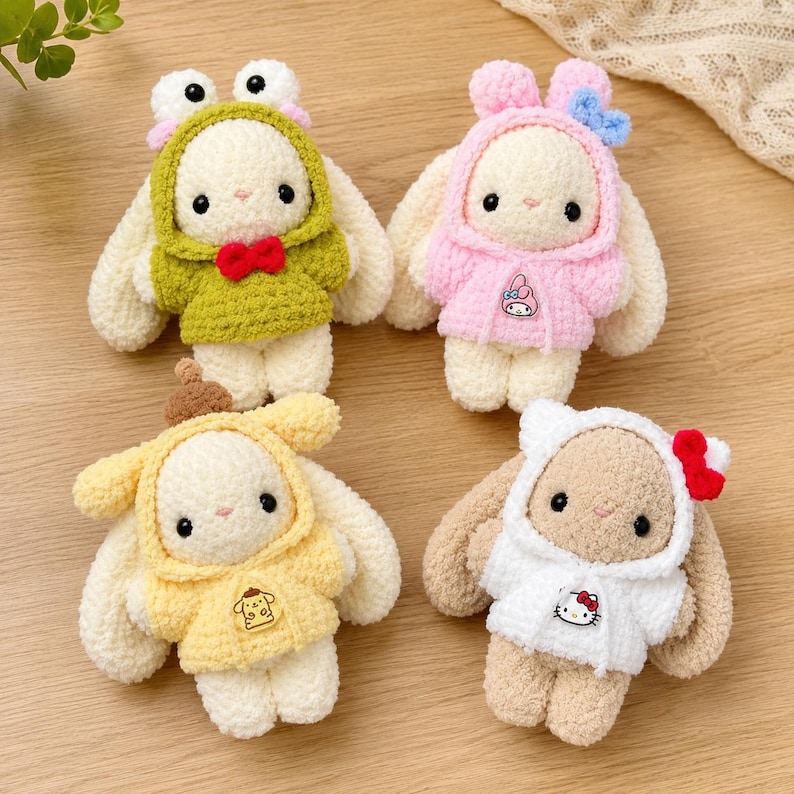 May include: Four plush crochet rabbit dolls wearing hoodies. One wears a green frog hoodie with a red bow, one a pink hoodie with a My Melody graphic, one a yellow hoodie with a Pompompurin graphic, and one a white hoodie with a Hello Kitty graphic.