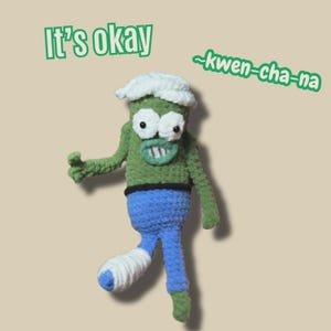 Può includere: Bambola mostro verde all'uncinetto con cappello bianco, pantaloni blu e gamba fasciata. La bambola ha un ampio sorriso e il testo "It's okay" e "~kwen-cha-na" in verde.