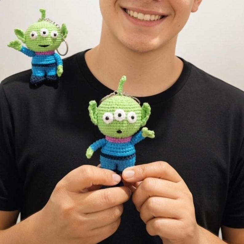 May include: Two green crochet alien keychains with three eyes, wearing blue and pink outfits. One keychain is held in front of a person, while the other is attached to a silver keyring. The keychains are handmade.