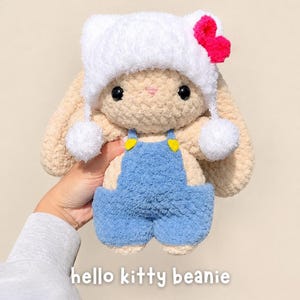 May include: A beige crochet plush rabbit wearing a white fuzzy beanie with a bright pink bow and two white pom-poms, dressed in blue overalls with yellow buttons. The text hello kitty beanie is printed at the bottom of the image.