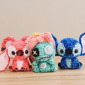 Pu&ograve; includere: Tre peluche fatti a mano all'uncinetto a forma di personaggio dei cartoni animati. I giocattoli sono nei toni del rosa, verde acqua e blu, con occhi neri a bottone e dettagli ricamati. I peluche sono appoggiati su una superficie di legno.