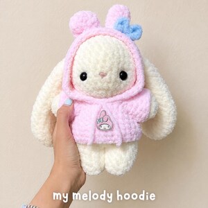 May include: A cream-colored plush rabbit toy with long ears, wearing a soft pink hooded sweatshirt. The hood features a small blue bow. A small My Melody character emblem is attached to the center of the pink hoodie. The toy has black eyes.