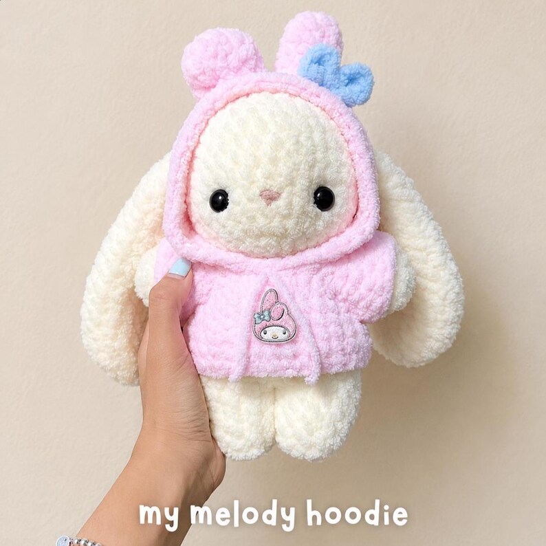 May include: A cream-colored plush rabbit toy with long ears, wearing a soft pink hooded sweatshirt. The hood features a small blue bow. A small My Melody character emblem is attached to the center of the pink hoodie. The toy has black eyes.