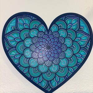 May include: A heart-shaped wooden artwork with a mandala design. The mandala features concentric patterns in teal, turquoise, and purple, set against a dark blue border. The artwork is mounted on a white wall.